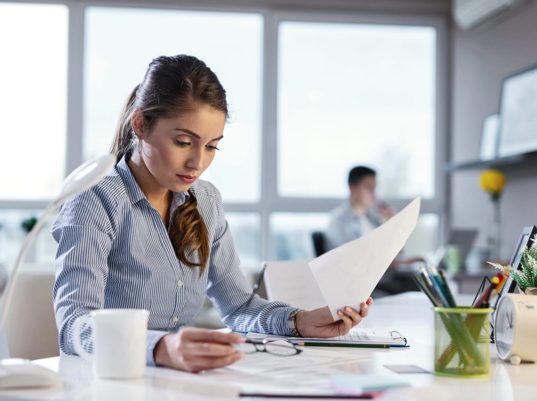 Young businesswoman reading business reports while doing paperwork at office desk. Her colleague is in the background.